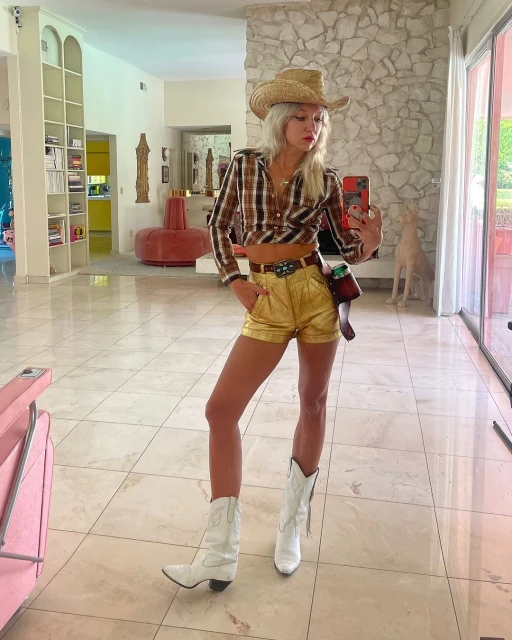 Woman in straw cowboy hat, plaid shirt, gold shorts, and white boots poses in a bright, airy living room taking a selfie.