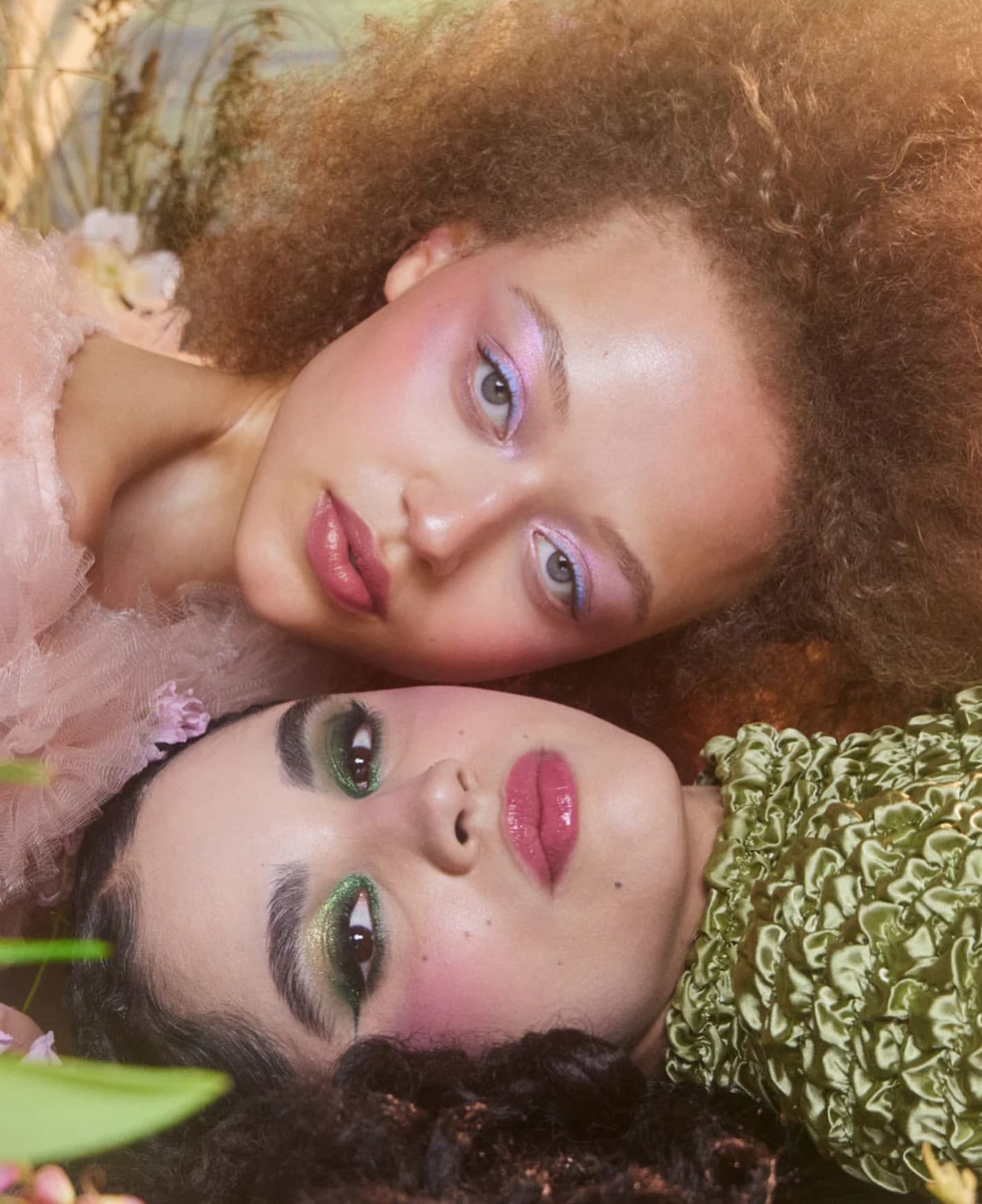 Two women with dramatic makeup lying together in a floral setting, eye-level angle, beauty and fashion photography, soft focus.