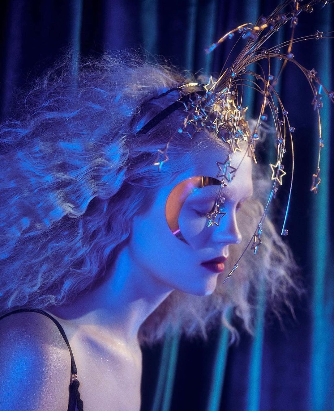Profile shot of a woman with fair skin, curly blonde hair, and an elaborate gold headpiece with star and moon details, moody blue lighting.