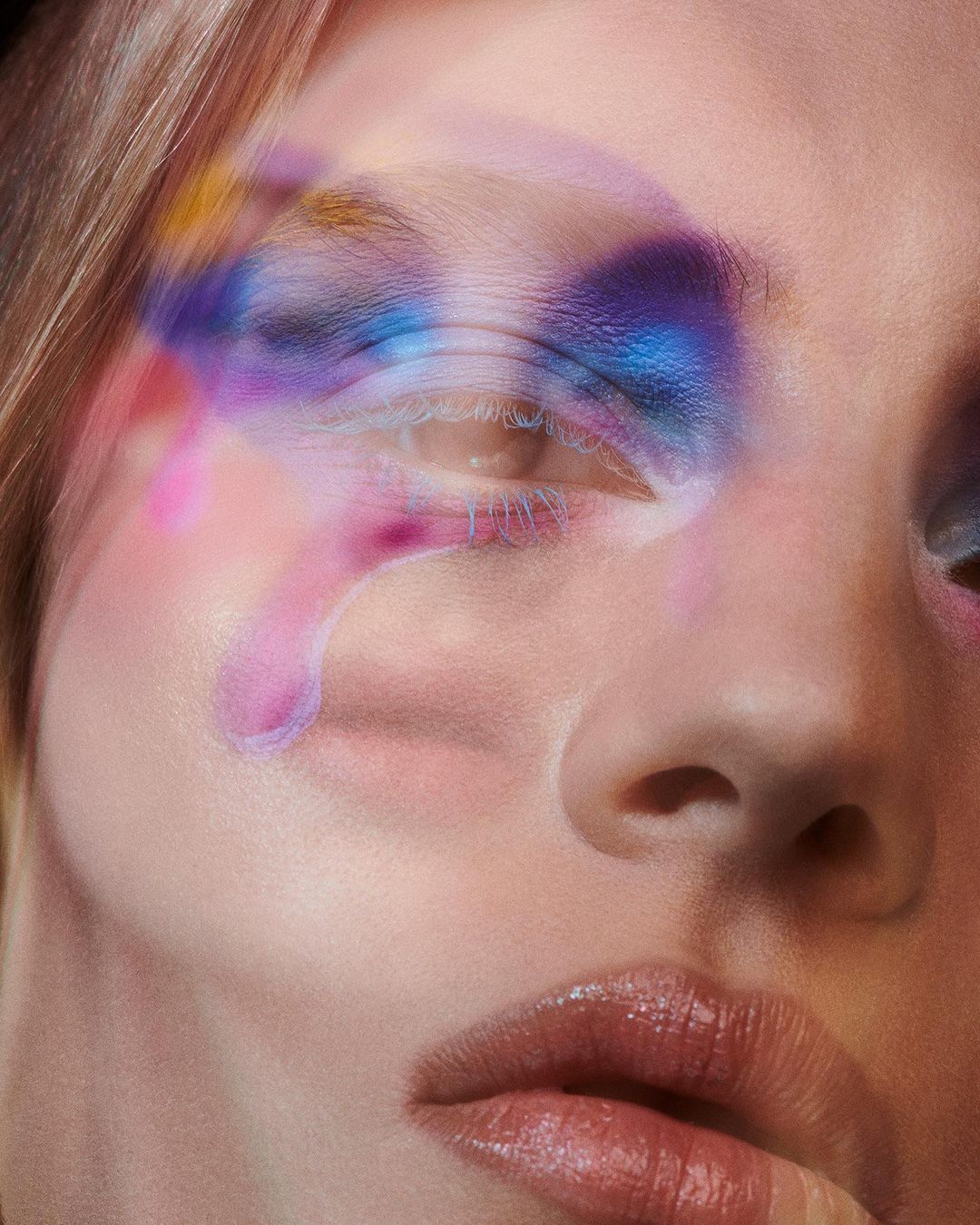 Close-up of a woman's face with artistic makeup in shades of blue, purple, and pink, creating a visually striking, blurred effect.