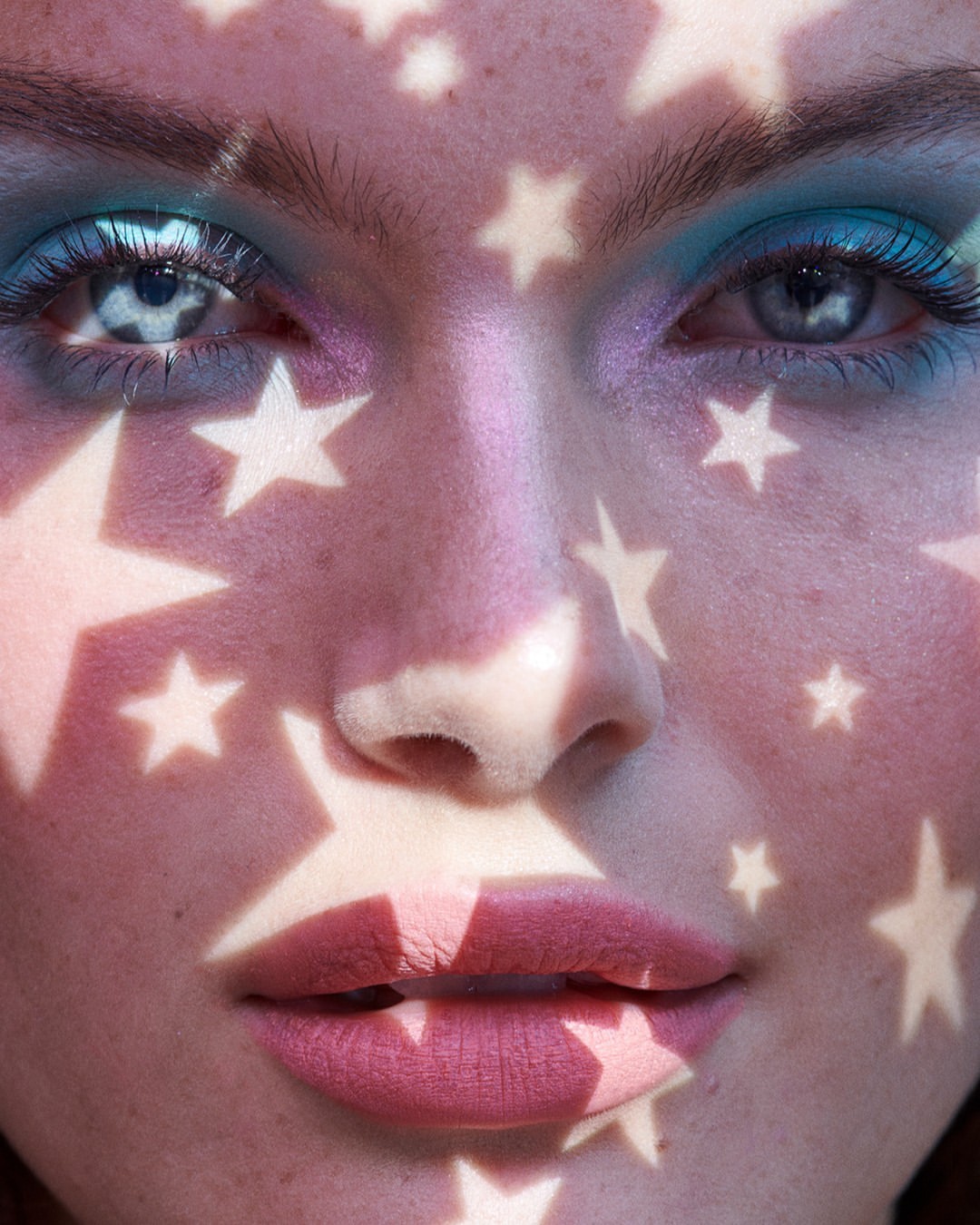 Close-up of a woman's face with vibrant makeup, featuring projected star shapes creating a unique and artistic visual effect.