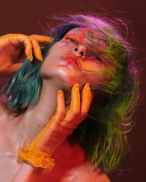 Close-up portrait of a person with brightly colored makeup and hair, wearing orange translucent gloves against a brown backdrop.