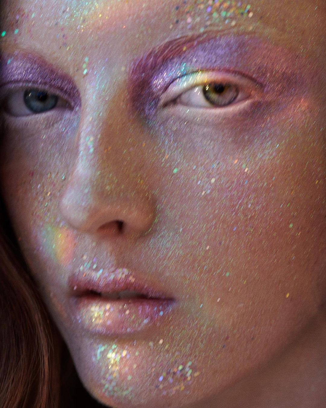 Closeup of a person's face with iridescent glitter makeup on the skin and eyelids. One eye is blue, and the other is green.