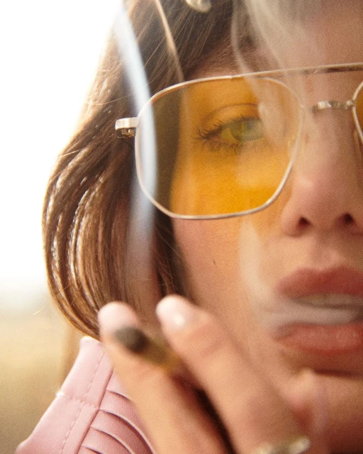 Close-up of a woman with yellow glasses smoking, showcasing hair, facial features, and fashion details against a bright background.