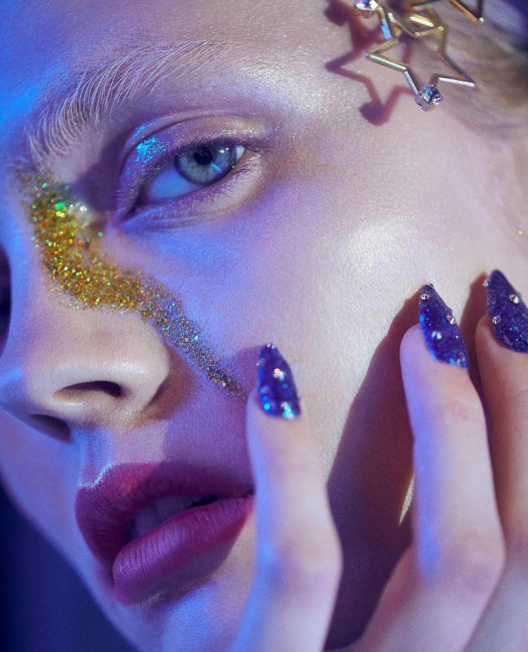 Close-up of a woman's face with gold glitter on her cheek and jeweled fingernails, accented by blue lighting and star-shaped hair clips.