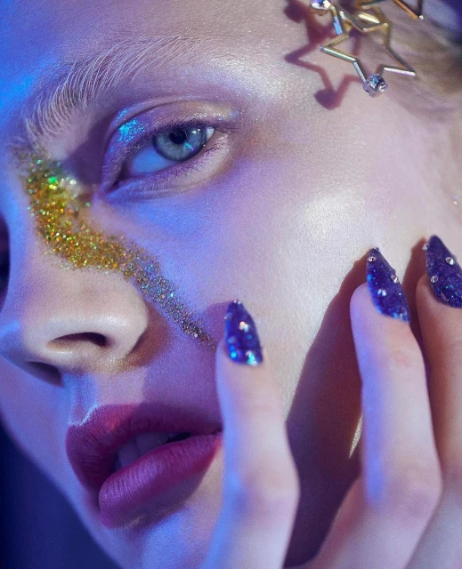 Close-up of a woman's face with gold glitter on her cheek and jeweled fingernails, accented by blue lighting and star-shaped hair clips.