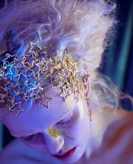 Close-up portrait of a woman with star headpiece and gold glitter eye makeup against a gradient blue background in a beauty shot.
