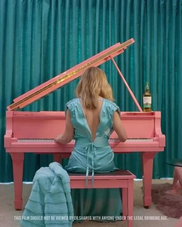 Woman in a turquoise dress playing a pink grand piano, back view with a teal curtain in the background, creating a vibrant color palette.