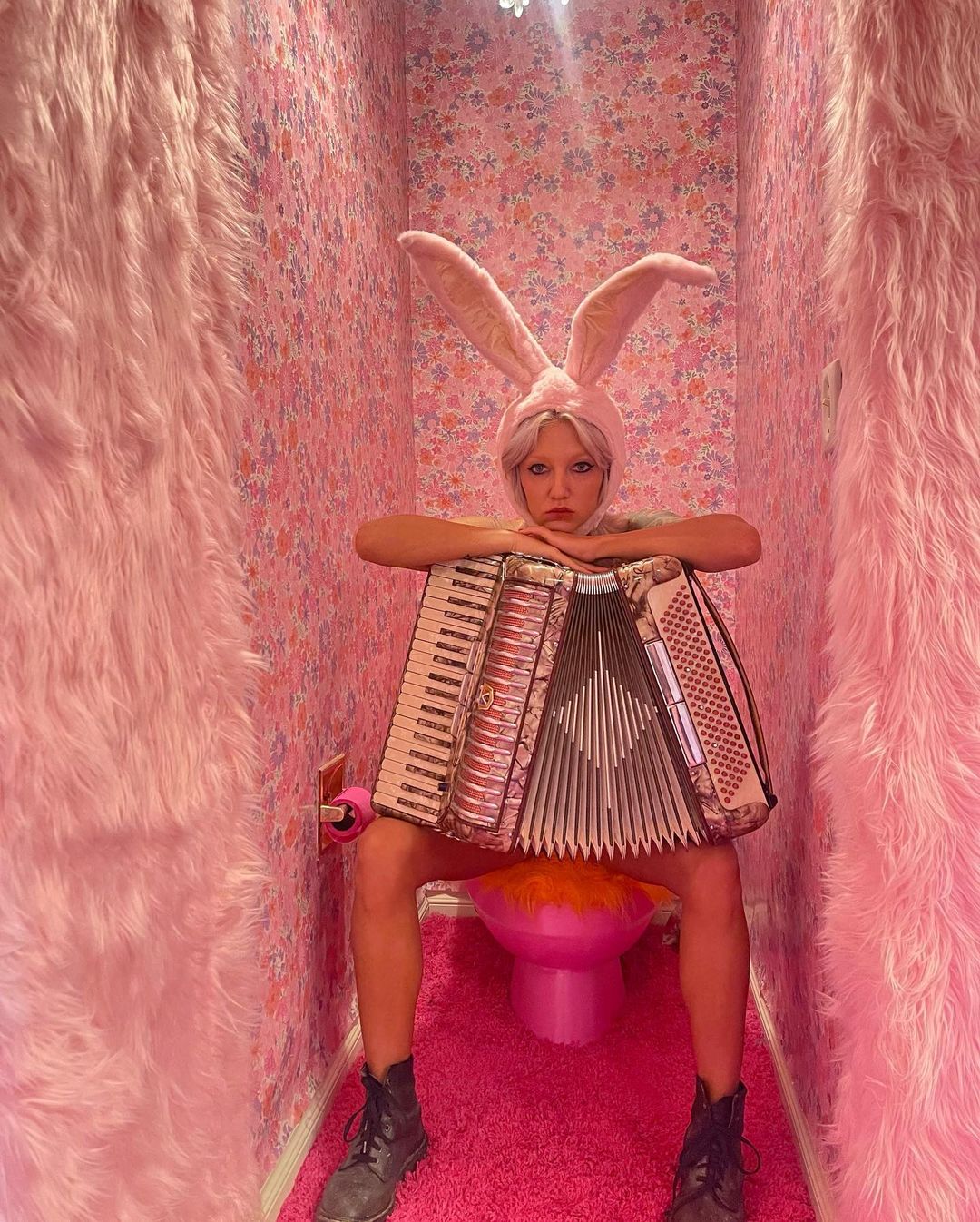 Woman in bunny ears sits on pink toilet holding accordion in pink room with floral wallpaper and faux fur wall coverings for a surreal effect.