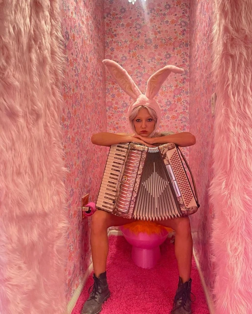 Woman in bunny ears sits on pink toilet holding accordion in pink room with floral wallpaper and faux fur wall coverings for a surreal effect.