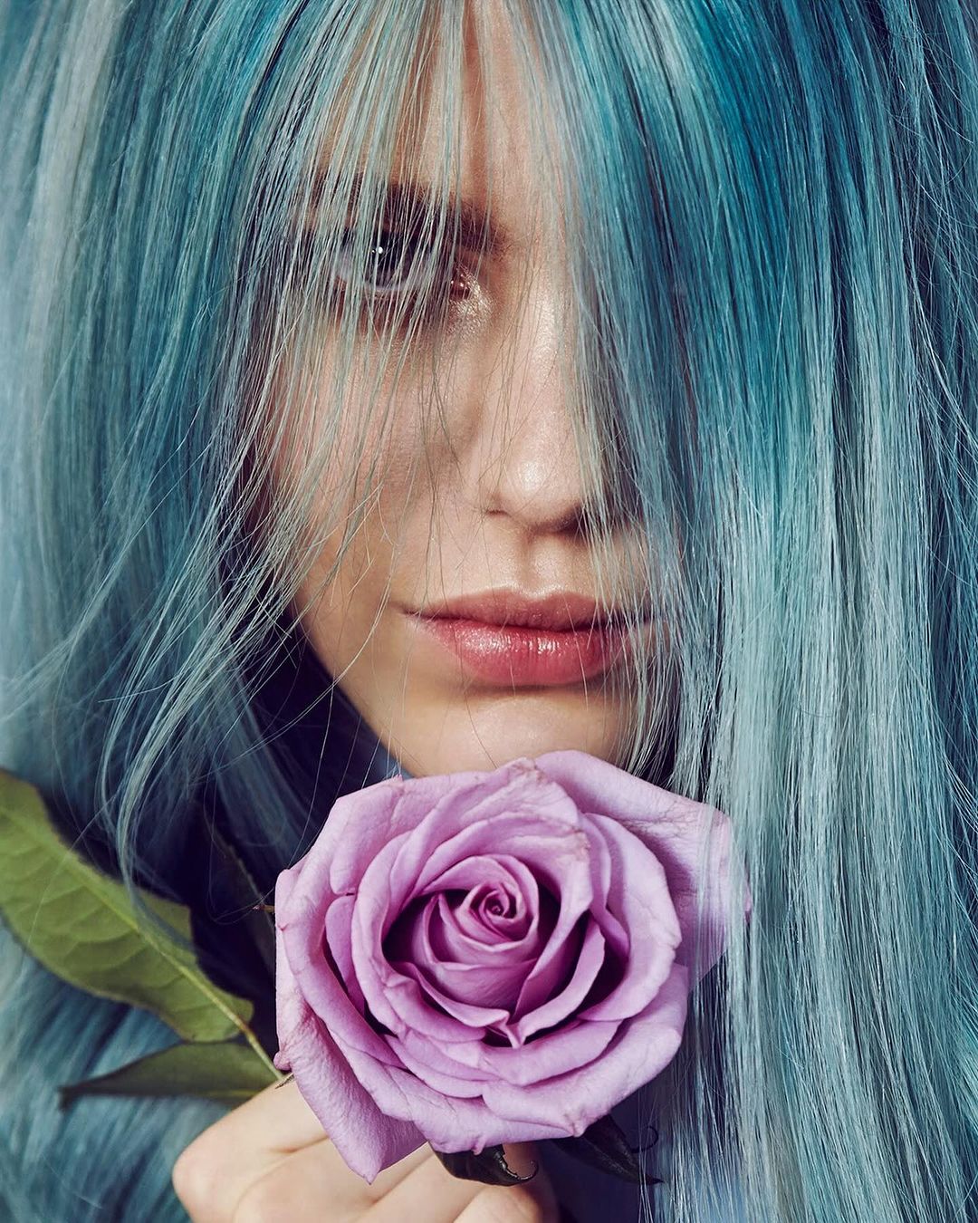 Close-up portrait of a woman with blue-toned hair holding a purple rose, focusing on her face and the flower against a muted background.