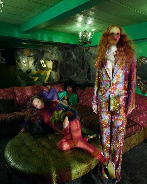 Three models in retro-inspired fashion pose in an indoor setting with a couch, green ceiling, and stone wall feature. 