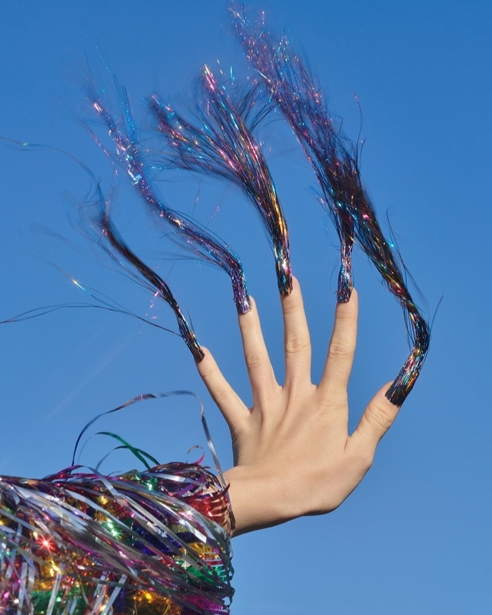 Woman's hand with long, glittery black nails outstretched against a clear blue sky, wearing a colourful metallic fringe sleeve.