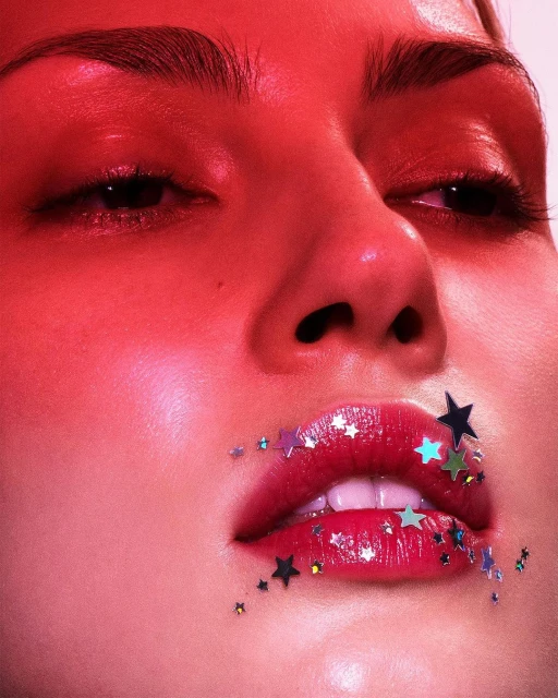Close-up beauty shot of a fair-skinned young adult female with pink lipstick and star-shaped glitter around her mouth in red light.