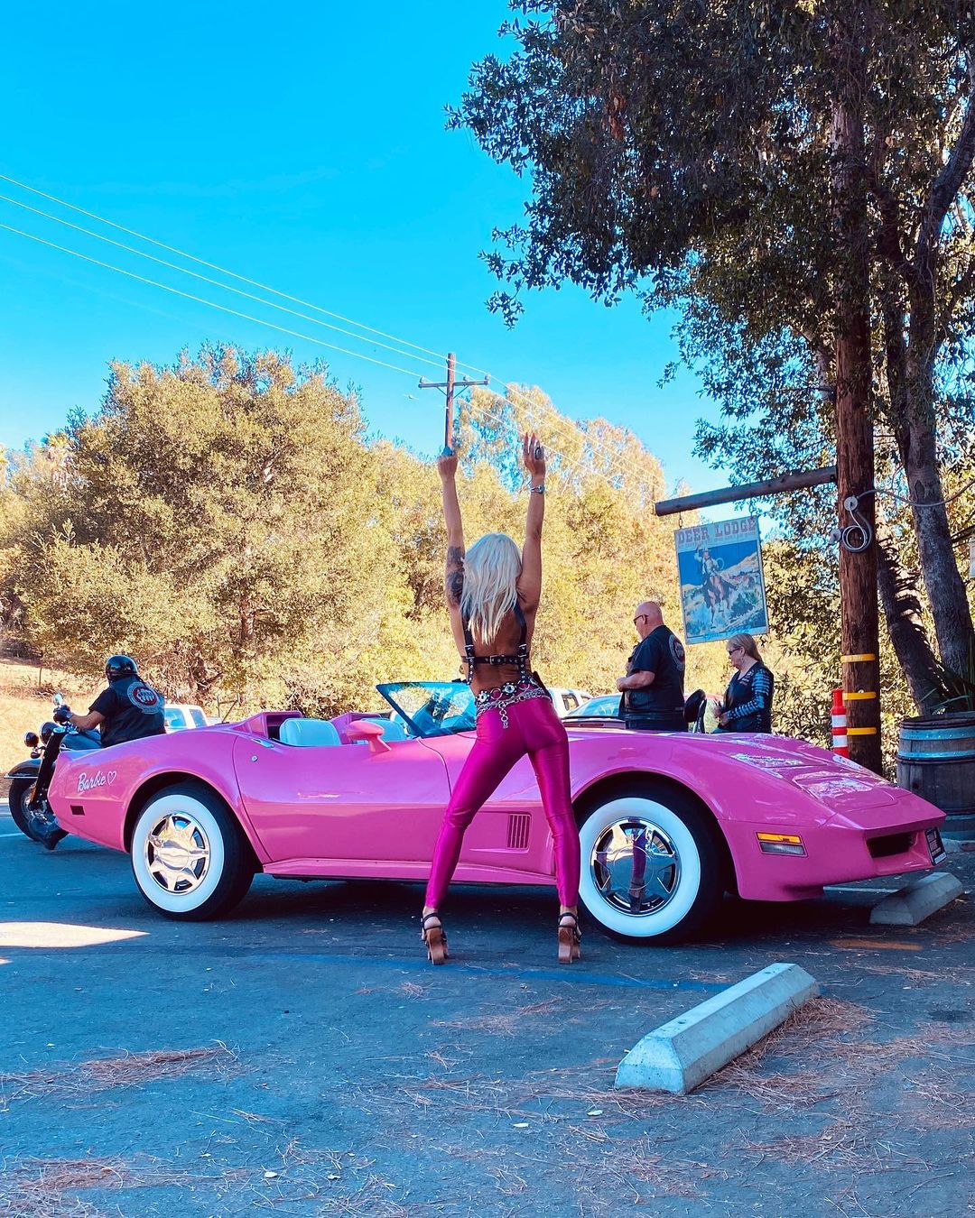 Blonde adult female stands by a pink convertible car with arms raised outdoors on a sunny day in a casual setting.