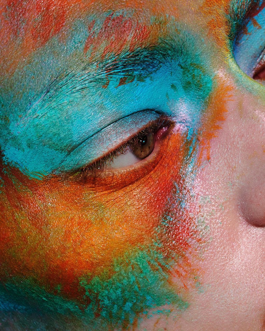 Close-up of a person's face with orange, blue, and red paint on the skin around the eye and eyebrow in a beauty-themed artistic style.