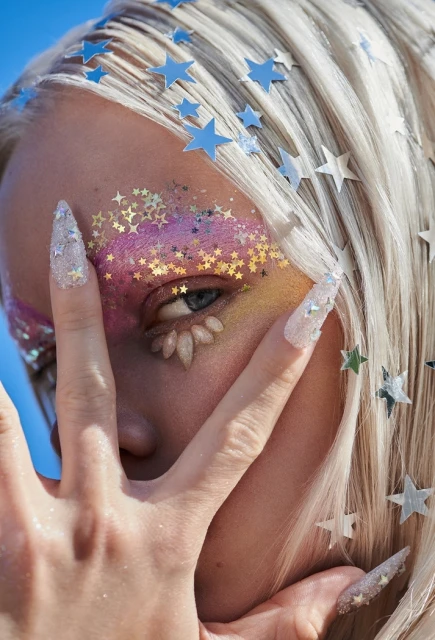 Woman with star-shaped decorations on platinum blonde hair, colorful glitter makeup, and stiletto nails poses against a blue sky.