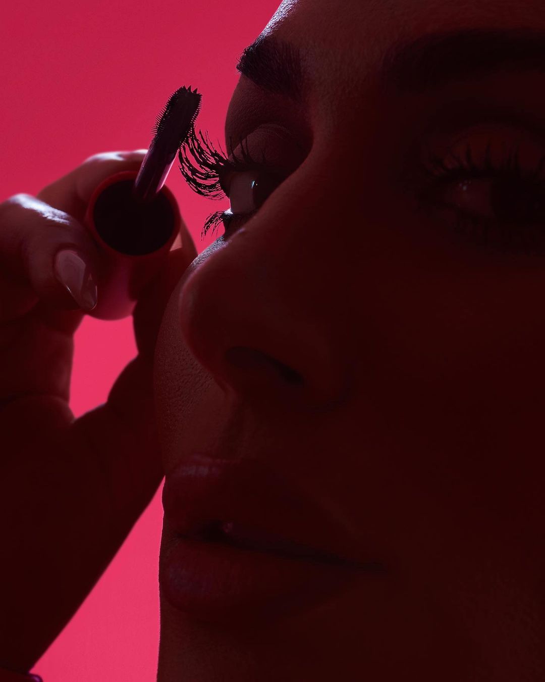 A close-up shot of a woman applying mascara, focusing on the eyelashes and the makeup wand against a bright pink background.