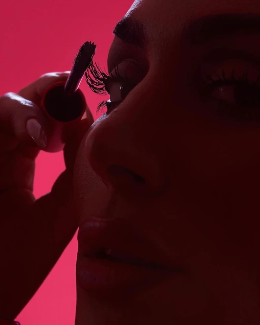 A close-up shot of a woman applying mascara, focusing on the eyelashes and the makeup wand against a bright pink background.