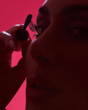 A close-up shot of a woman applying mascara, focusing on the eyelashes and the makeup wand against a bright pink background.