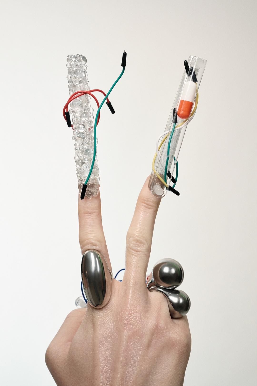 Close-up of a hand in a V-sign with jewelry, decorative fingernails, and attached wires and capsules against a neutral background.