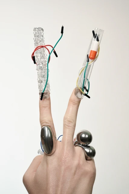 Close-up of a hand in a V-sign with jewelry, decorative fingernails, and attached wires and capsules against a neutral background.