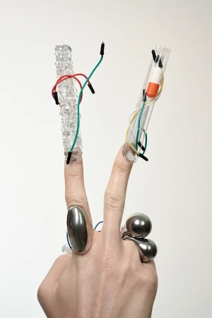 Close-up of a hand in a V-sign with jewelry, decorative fingernails, and attached wires and capsules against a neutral background.