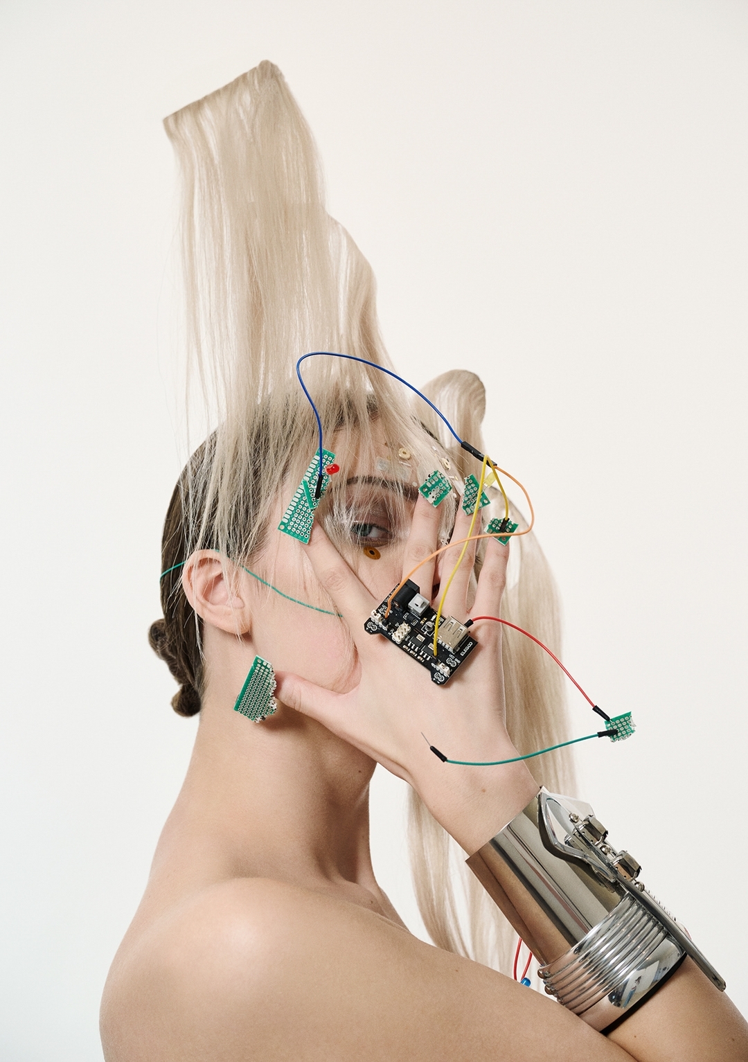 Artistic portrait of a woman with platinum blonde hair, electronic components, and a metallic arm cuff against a white backdrop.