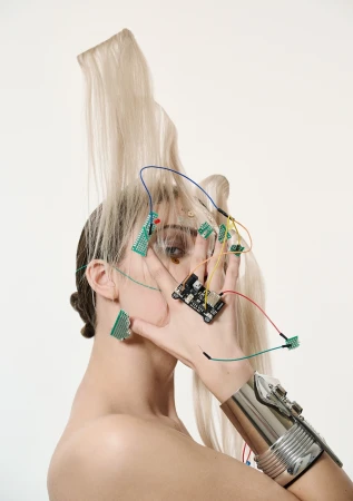 Artistic portrait of a woman with platinum blonde hair, electronic components, and a metallic arm cuff against a white backdrop.