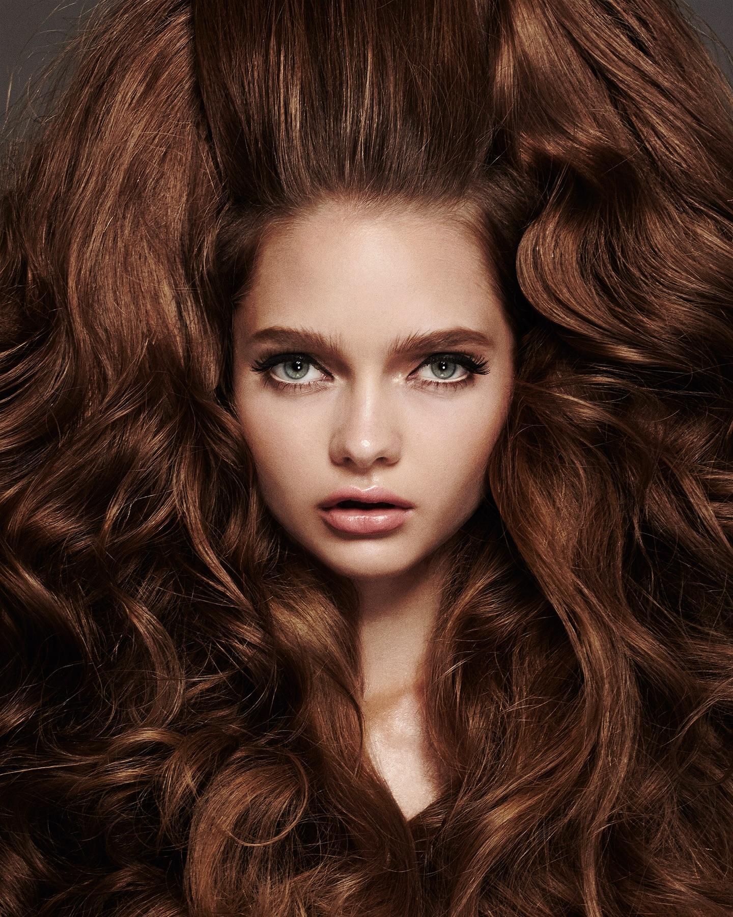 Close-up portrait of a young woman with voluminous auburn, wavy hair framing her face; makeup with defined eyes and full lips.