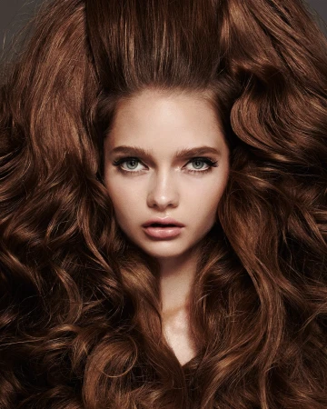 Close-up portrait of a young woman with voluminous auburn, wavy hair framing her face; makeup with defined eyes and full lips.