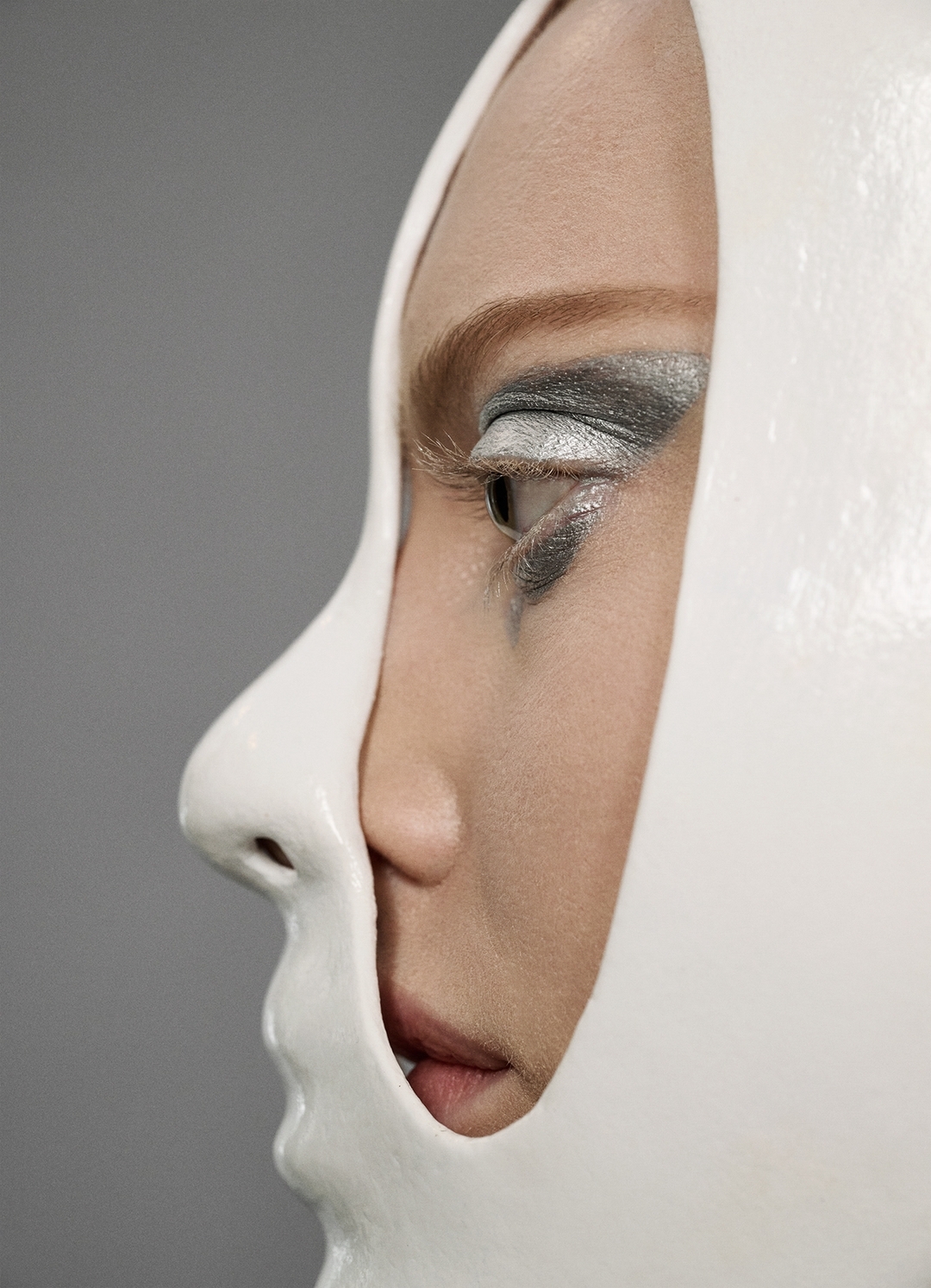 Profile of a woman with creative makeup and a partial white mask on a gray background, artistic eye makeup, modern beauty concept.