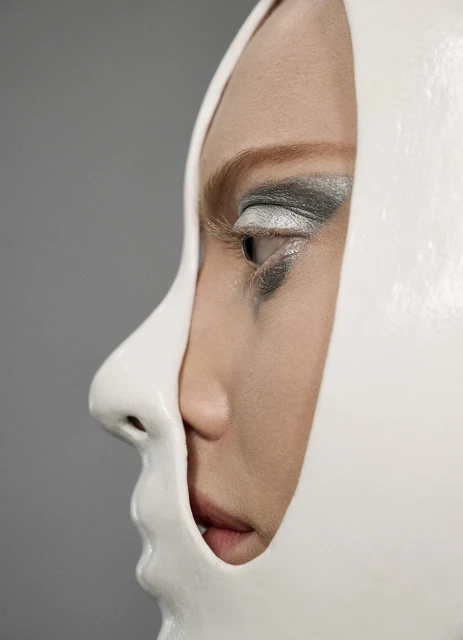 Profile of a woman with creative makeup and a partial white mask on a gray background, artistic eye makeup, modern beauty concept.