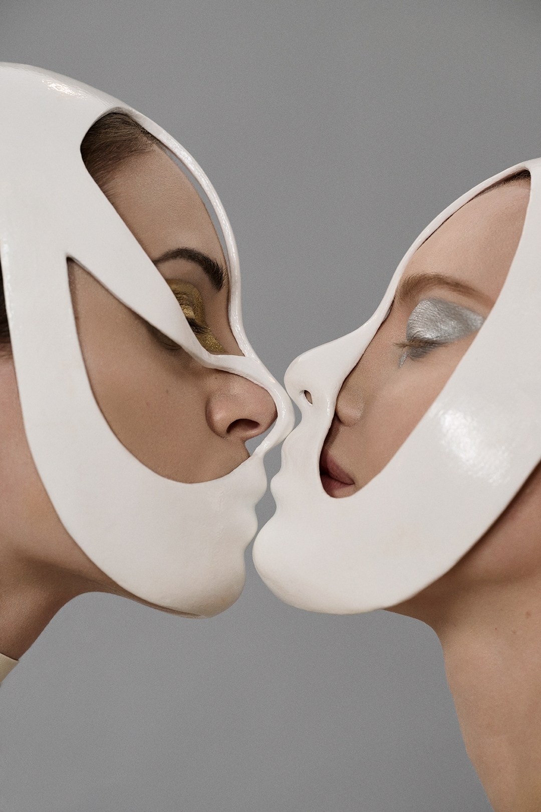 Close-up of two people with light skin wearing sculptural face masks, facing each other in a studio setting with a neutral gray background.