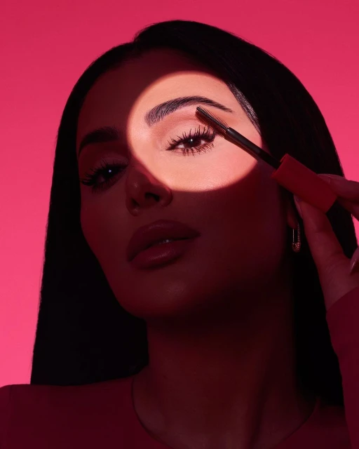 Close-up studio shot of a woman applying eyebrow gel with a wand in a brightly lit spotlight against a vibrant pink backdrop.