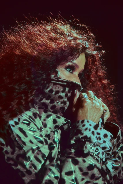 A woman with red hair and striking green eyes wears a leopard-print jacket, her hands near her face in a dimly lit setting.