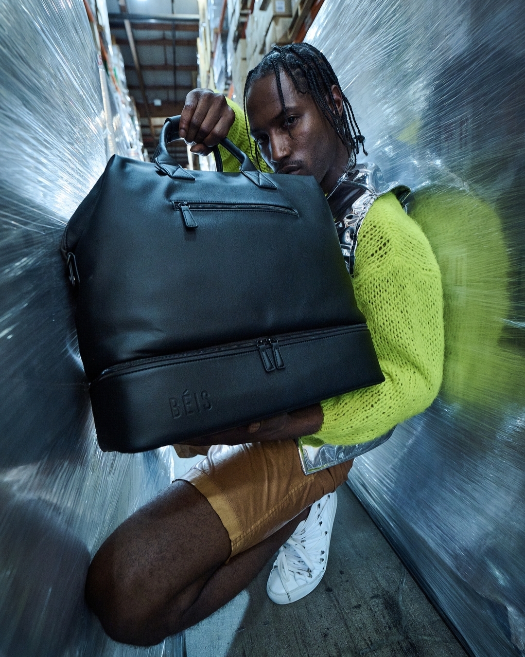 Person crouches in warehouse holding black bag with 'BÉIS' logo, wearing yellow sweater, silver vest, and tan shorts, low angle.