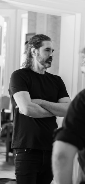 Black-and-white medium shot of an adult man with long hair in a ponytail, arms crossed, wearing a black shirt in an indoor setting.