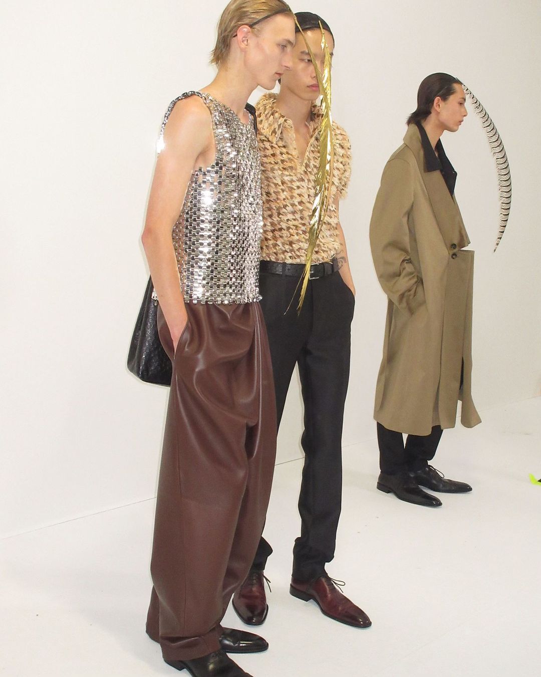 Three fashion models pose in a photo studio wearing shiny shirts, brown pants, and a trench coat, creating a modern editorial shot.