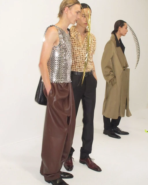 Three fashion models pose in a photo studio wearing shiny shirts, brown pants, and a trench coat, creating a modern editorial shot.