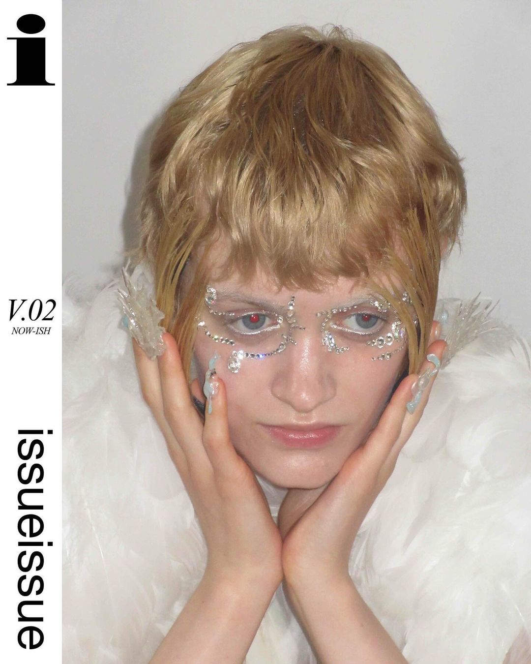 Close-up portrait of a blond adult woman with bejeweled face, hands on face, wearing a feathered boa in a white studio setting.