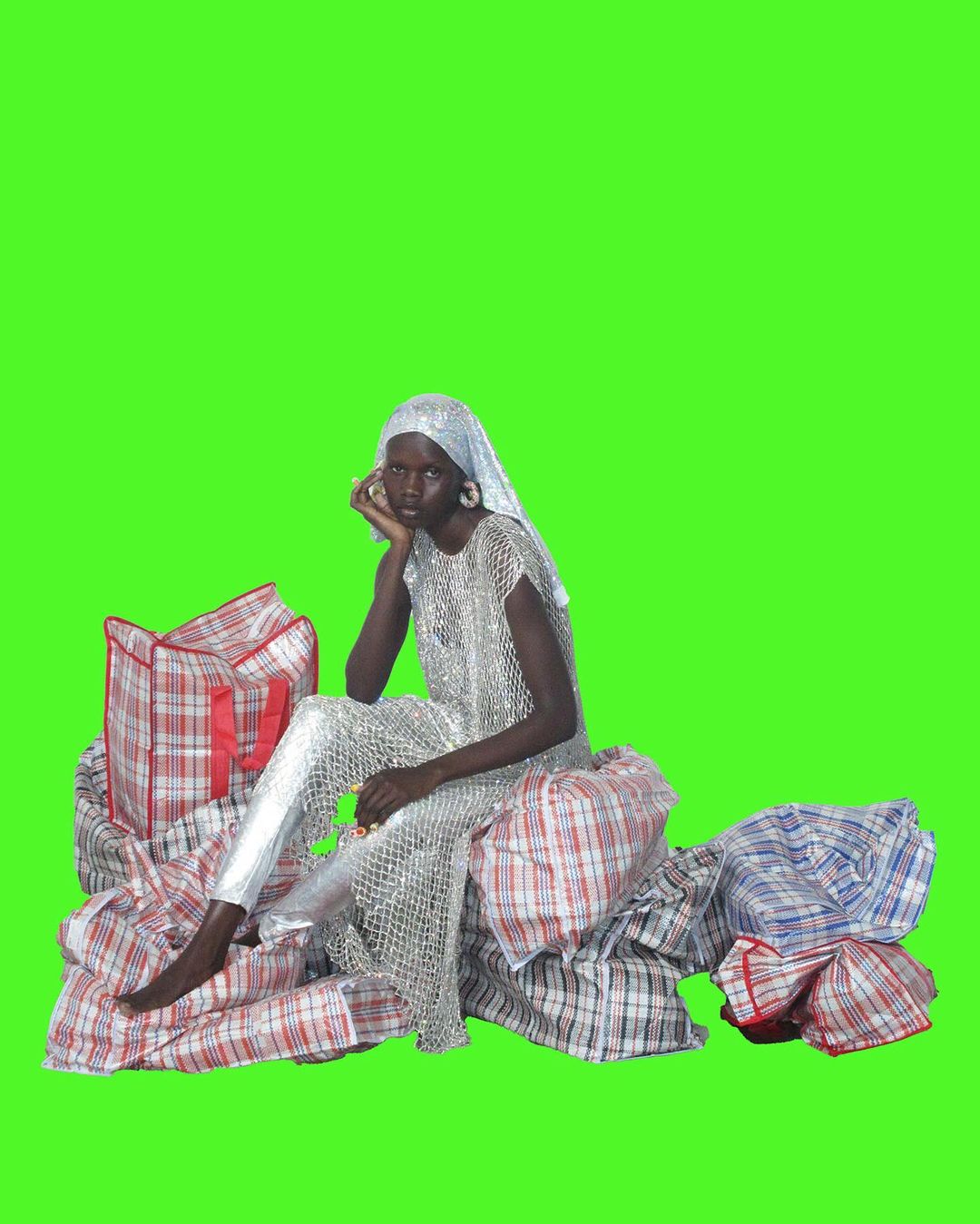 A dark-skinned model in shimmering silver top and pants poses on a pile of plaid shopping bags against a vibrant green background.