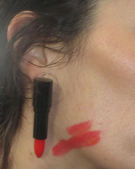 Close-up of a fair-skinned adult's face, showcasing lipstick applied on the skin and a lipstick tube held to the cheek by the ear.