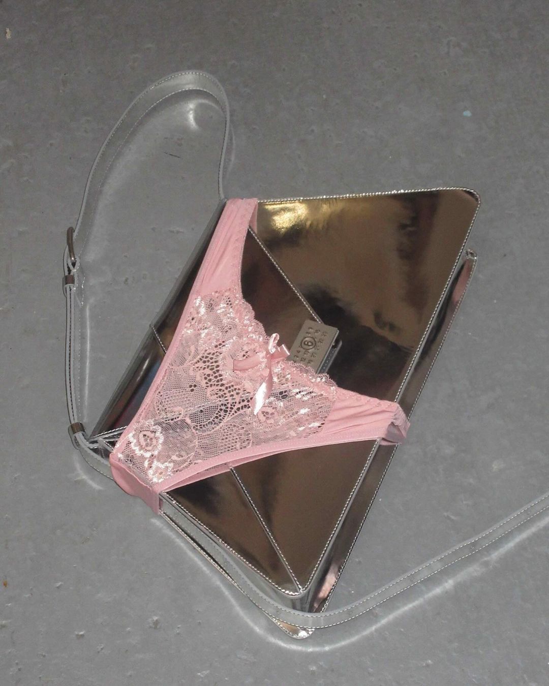 Metallic silver envelope clutch handbag accessorized with delicate, light-pink, floral-lace lingerie on a gray concrete surface.