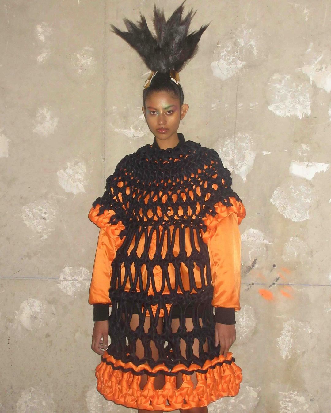 Woman with dramatic feathered hairstyle and orange-trimmed black netting dress stands against a mottled gray wall in a fashion-forward pose.