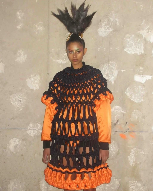 Woman with dramatic feathered hairstyle and orange-trimmed black netting dress stands against a mottled gray wall in a fashion-forward pose.