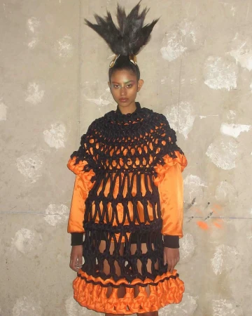 Woman with dramatic feathered hairstyle and orange-trimmed black netting dress stands against a mottled gray wall in a fashion-forward pose.