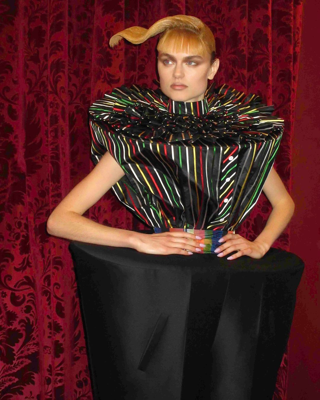 Fashion model poses in a striking, sculptural dress with a striped bodice and black skirt against a dark red, patterned backdrop.