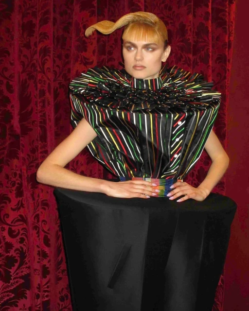 Fashion model poses in a striking, sculptural dress with a striped bodice and black skirt against a dark red, patterned backdrop.