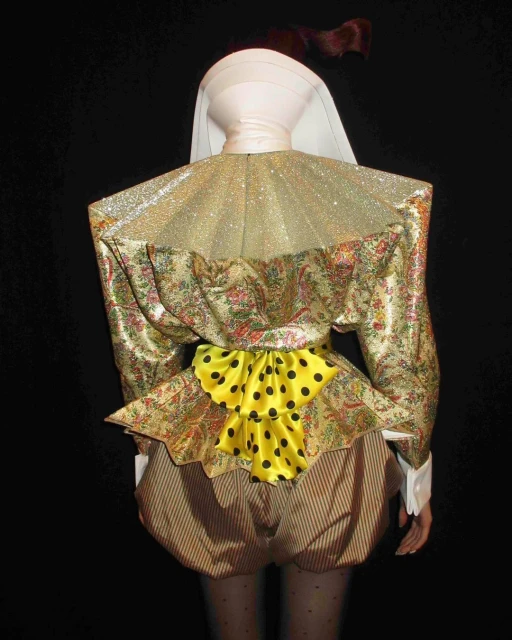 Costumed mannequin from behind wears gold floral jacket with pleated collar and yellow bow, with striped shorts, against black backdrop.